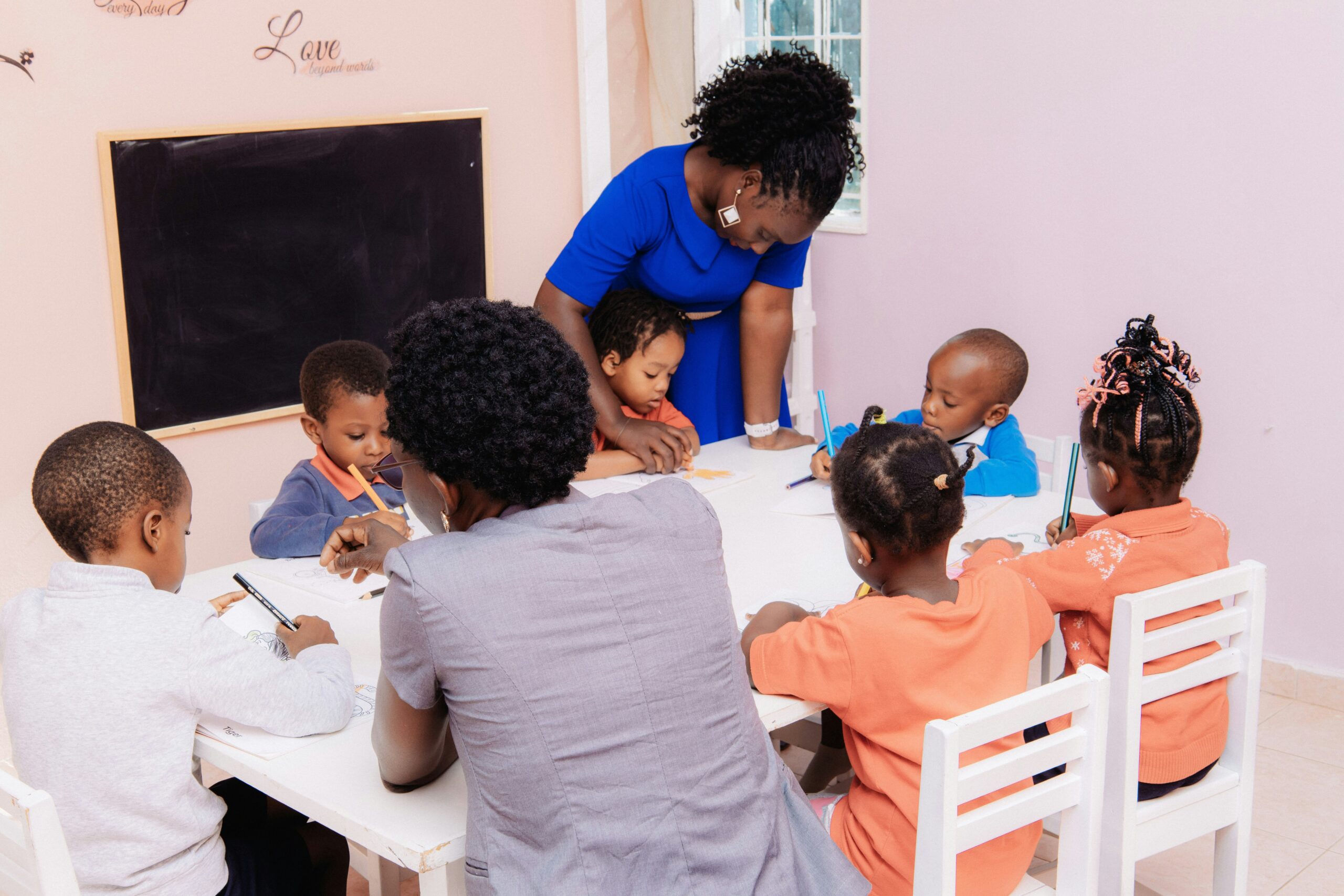 A teacher helps young children draw together at a brightly lit classroom table