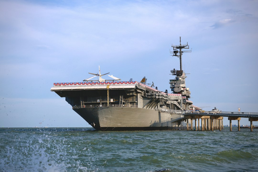 Large aircraft carrier docked beside blue water under a bright sky, illustrating the expanded U.S. Middle East deployment