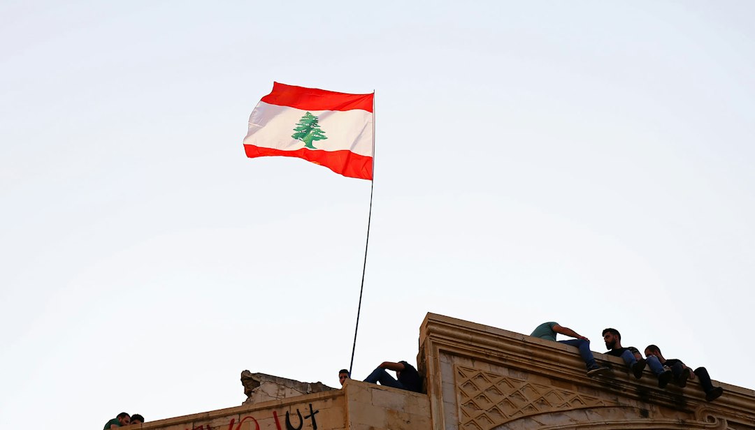 Lebanon’s red and white national flag with its green cedar flying above Beirut in bright daylight, representing the country’s humanitarian crisis.